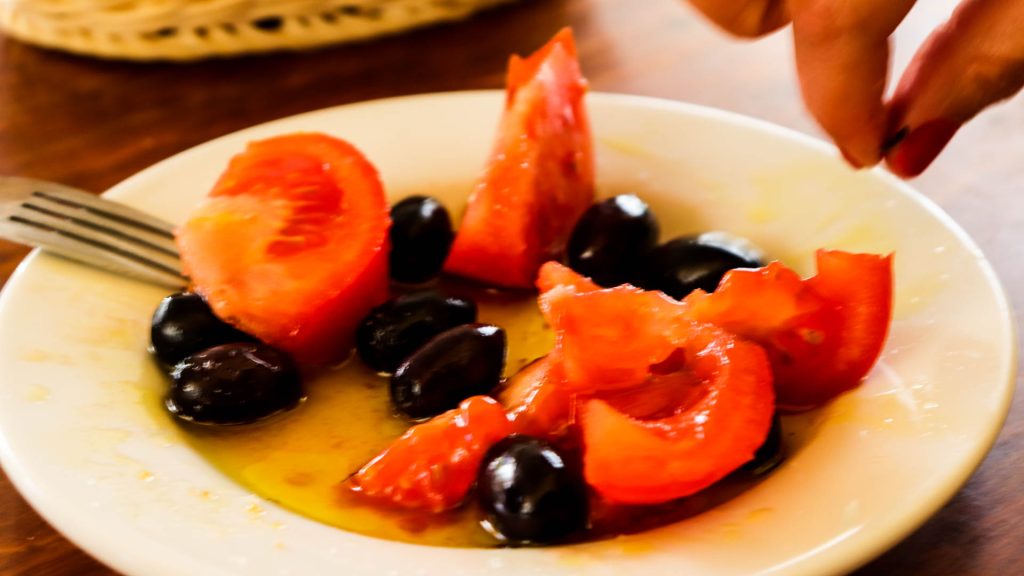 A rustic outdoor table in a sunny Cretan olive grove, laden with traditional farm-to-table dishes like dakos, wild greens, local cheeses, and a carafe of extra virgin olive oil, with ancient olive trees in the background.