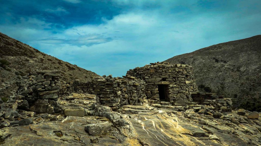A circular dry-stone shepherd's hut (mitato) on the rocky Nida Plateau of Mount Psiloritis, with a clear blue sky and the rugged limestone peaks of the geopark in the background.